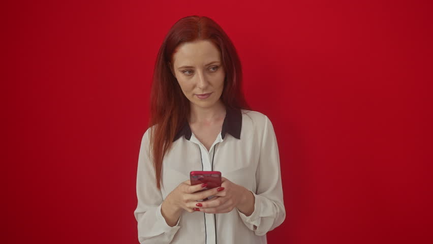 Young caucasian woman confidently holds smartphone, standing with serious expression in defense, gesturing stop sign with hand over isolated red background
