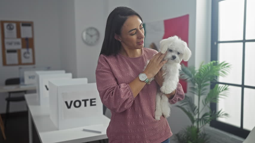 Hispanic woman holding a bichon maltes dog in an indoor college polling station with a banner.