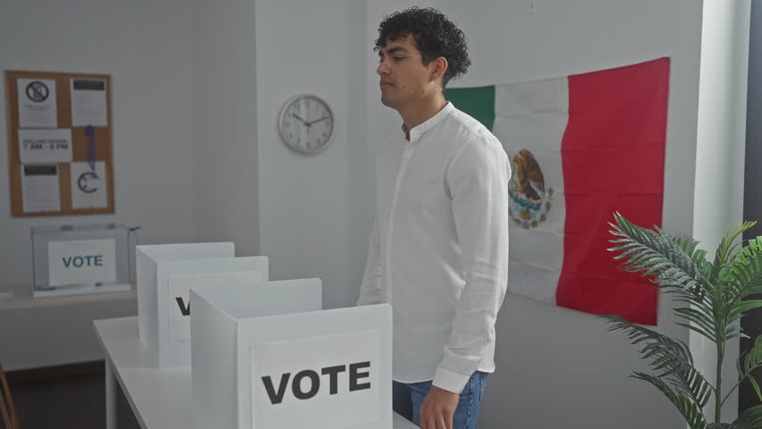 A young hispanic man gives a thumbs up in a mexican voting station with ballot booths and a flag in the background.