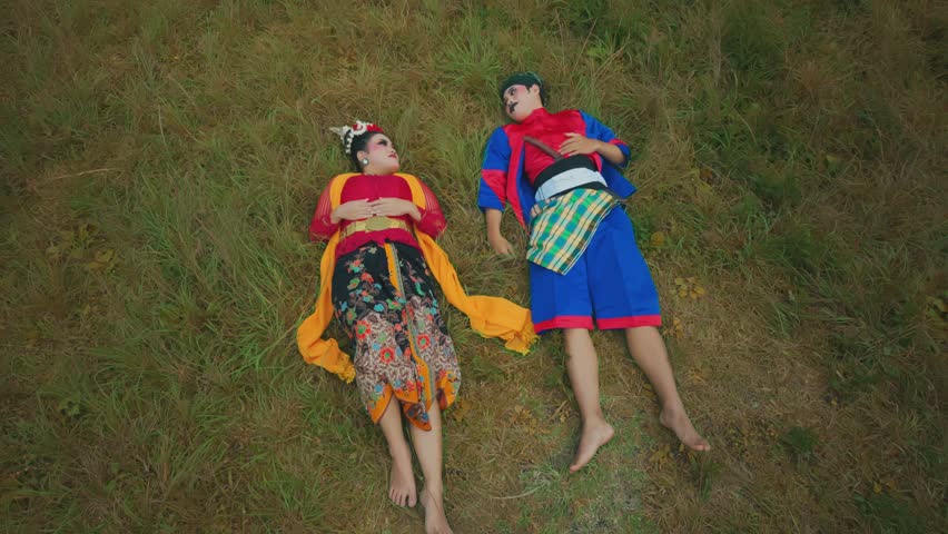 A man and a woman in traditional colorful clothing lying on grass, looking at each other.