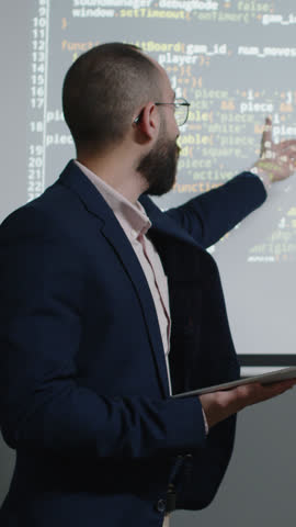 Medium vertical shot of young programming teacher or college professor in glasses, with tablet pointing to coding sequence on screen while explaining new subject to audience
