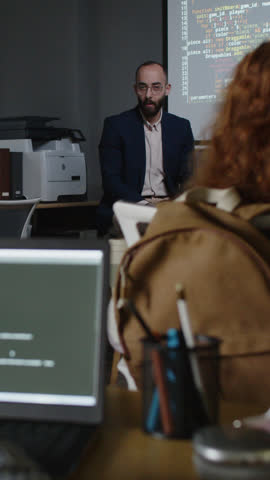Medium rear vertical shot of group of unrecognizable schoolchildren sitting at desks with laptops with computer code, listening to young male teacher speaking on new topic in front of projector screen