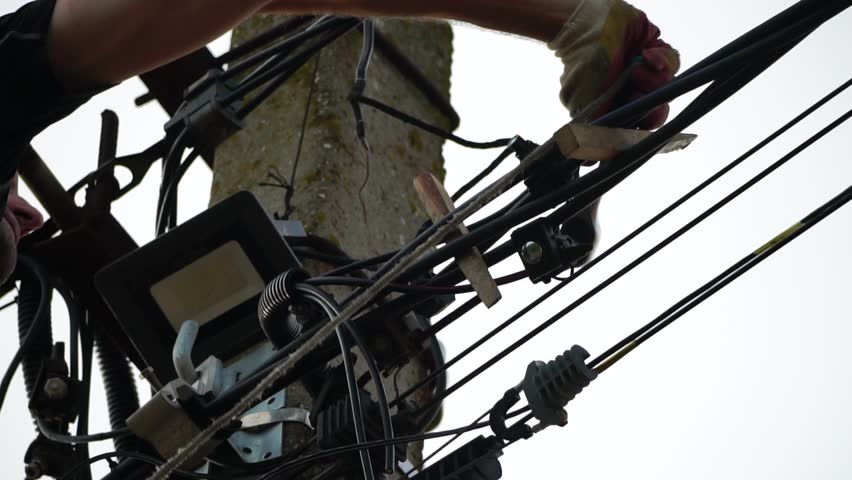 Skilled electrician fixes wires standing on ladder near high pole against blue sky on summer day backside view. Electrical service and mounting on the pole