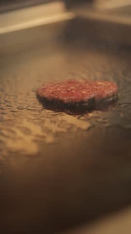 A closeup vertical footage of a chef's hand using a stainless spatula to flip burgers on a grill before adding the cheese, with blur background