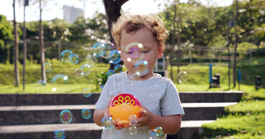 Toddler joyfully plays with small bubble machine, watching soap bubbles fly towards camera on breezy day. His eyes light up with imagination and wonder as he blows at bubbles