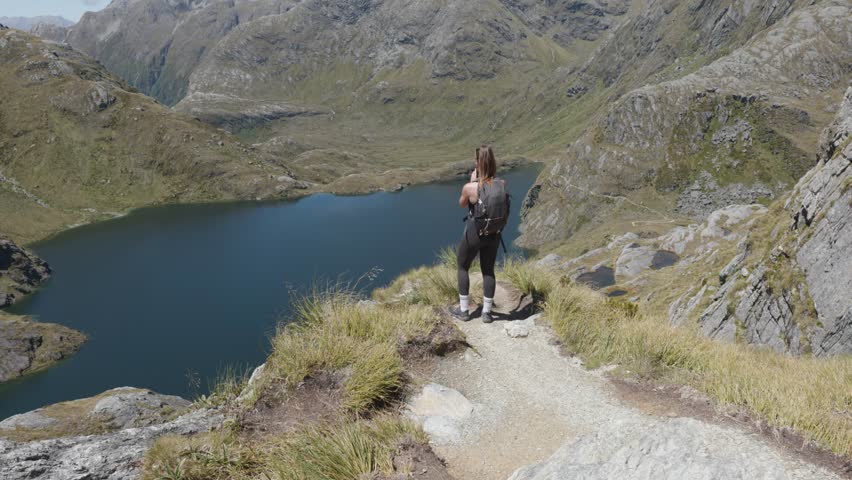 Young caucasian woman hiking and taking a video from a viewpoint of Harris Lake on a sunny summer day at Routeburn Track, New Zealand.
