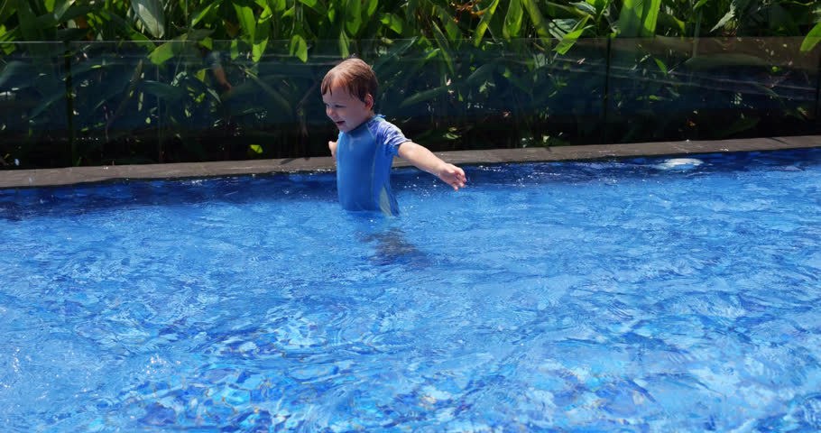 Joyful toddler stands in shallow wading pool, splashing water with his hands and jumping up and down, creating large splashes. On a sunny, hot day, baby boy energetically plays in water