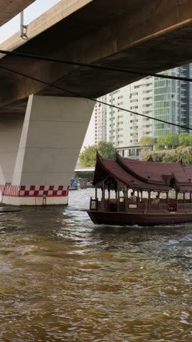 A vertical footage of traditional wooden Thai boat sailing on Chao Phraya River under a bridge in Bangkok, Thailand