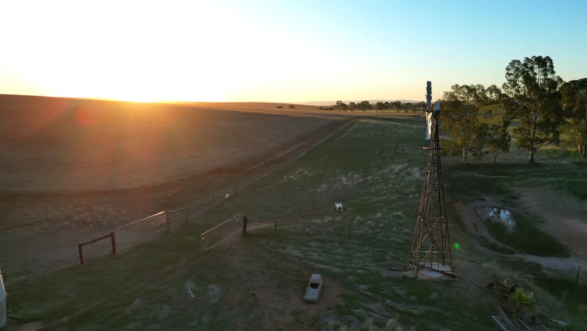 A drone footage of the sun setting over a rural farm with old farm-windmill and gum trees, Australia