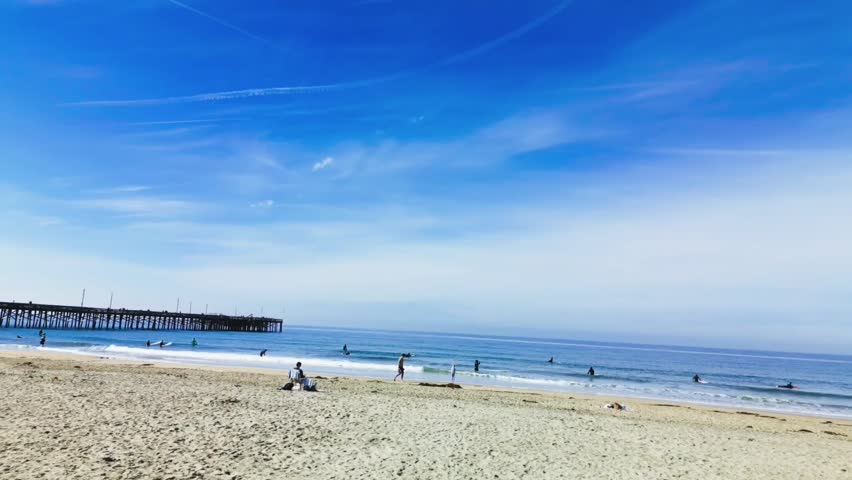 A drone footage of Huntington Beach in California on a sunny day with beachgoers swimming, relaxing on the sand and walking on pier