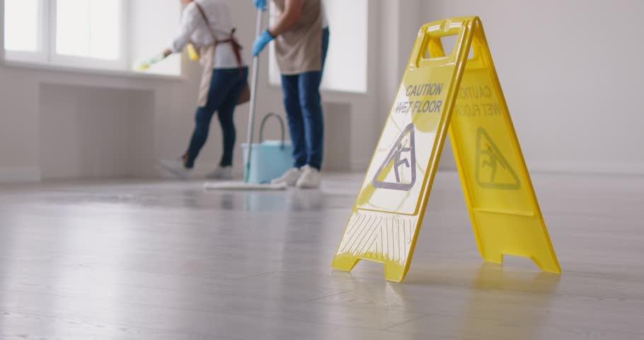 Close up shot of a Caution Wet Floor sign with cleaning service staff on background mopping floors in an office or home. This warning sign ensures safety during cleaning or household chores process.