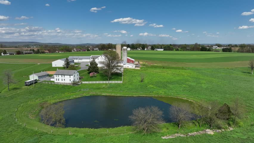 Aerial approaching shot of idyllic farm house with silo and private lake. Sunny day in american countryside during spring season. Stable and barn in PA, USA.