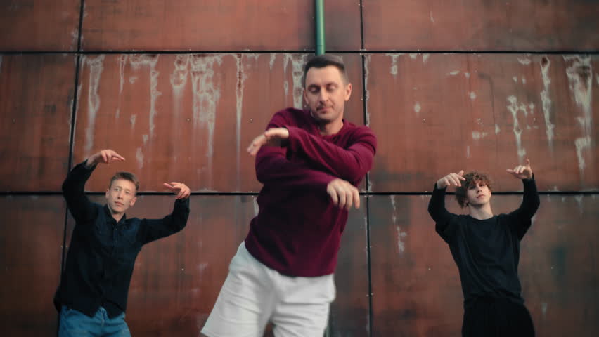 Middle-aged man breakdancer performing contemporary hip-hop choreography in city street, medium shot. Portrait of adult male dancer hangout out with group of teens, forever young and active person