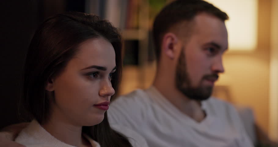 Hadsome man and attractive young woman sit in silence watching emotional drama back view reveals them wiping away tears and holding each other's hand for comfort selective focus close up shot.