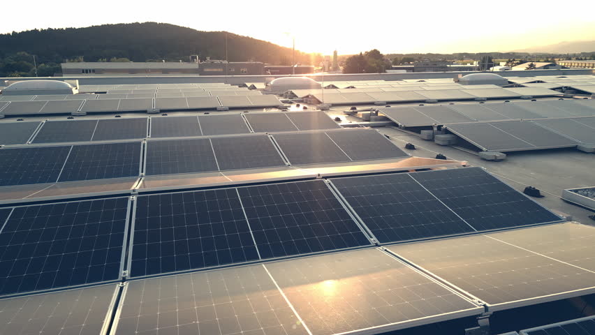Moving forward above rooftop solar panel array with sunset sun on horizon, aerial view. Photovoltaic modules on industrial building roof for green energy generation drone shot