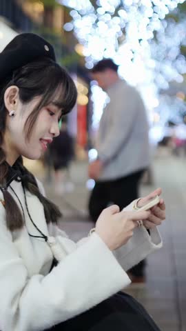 Vertical video of a young and cute Taiwanese woman in her 20s sitting and spending time amidst beautiful illuminations at night in a high-end shopping mall area of Xinyi District, Taipei, Taiwan