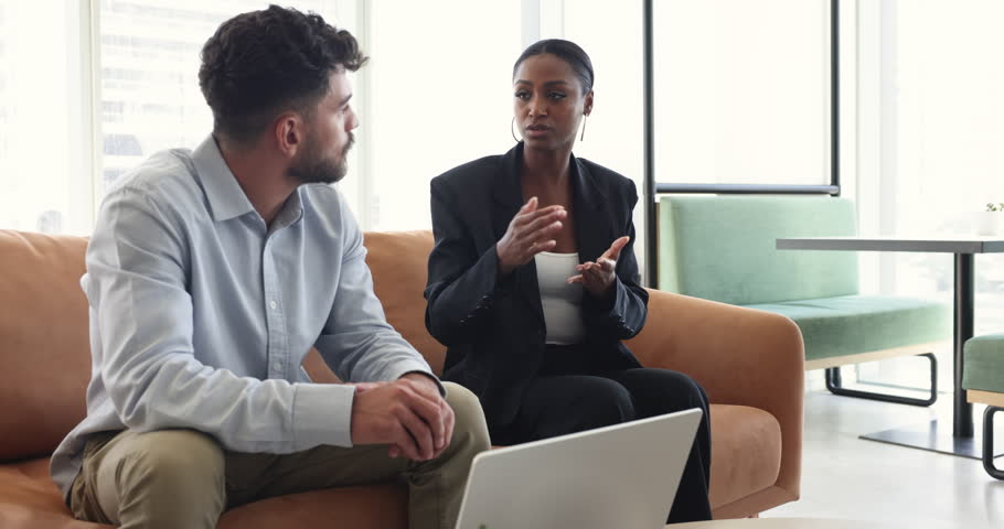 Multiethnic couple of office friends chatting in lobby, talking at laptop, discussing company life, job, career, getting cheerful, happy, sitting on couch. African employee woman speaking to colleague