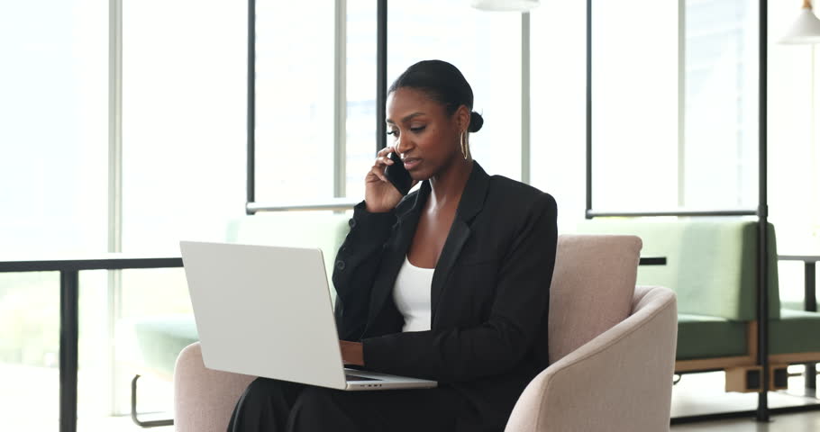 Cheerful beautiful African American business woman speaking on mobile phone at laptop, working from co-working cafe, talking on telephone call, using online app on computer for conversation