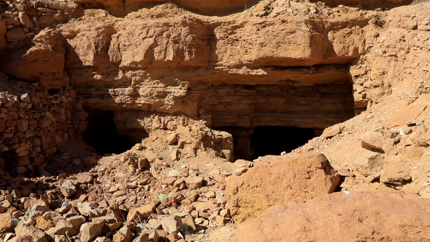 Ancient cave entrance in a rugged desert terrain with bright sunlight illuminating the rock formations