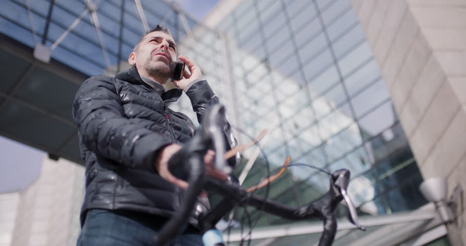 Urban professional takes a phone call outdoors, leaning on his bicycle with a contemporary cityscape in the background.