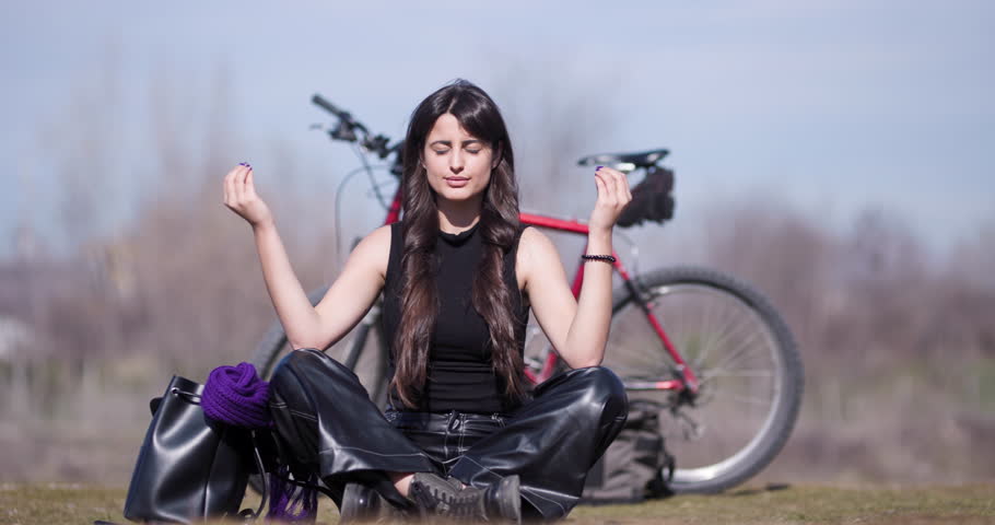 A young woman finds tranquility in meditation outdoors beside her mountain bike, surrounded by nature on a clear day.