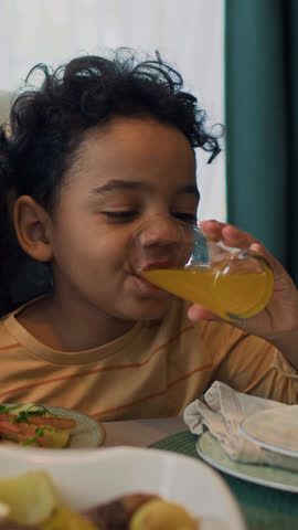 Medium close-up vertical shot of 5-year-old African American boy sitting at table at family dinner party, drinking glass of orange juice, and unrecognizable mother patting his shoulder