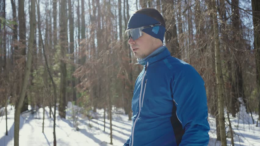 Male skier looks at watch checking time in winter forest. Man athlete in protective glasses engages in skiing session preparing for competition on sunny morning. Seasonal activity for sporty people