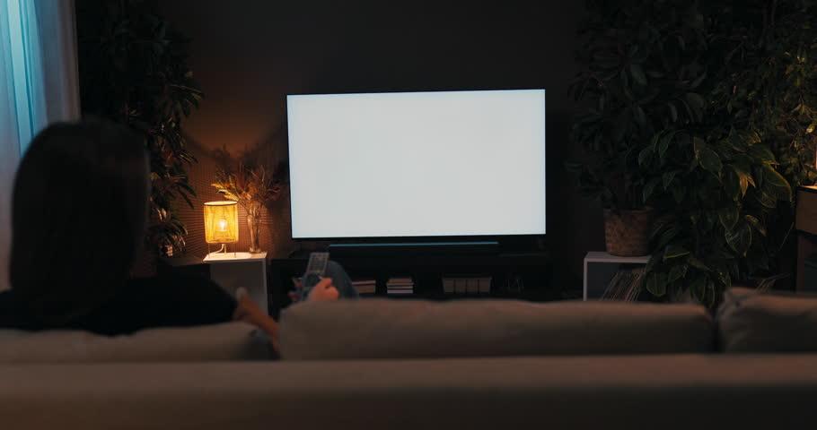 Relieved young brunette woman with long dark hair sitting in the living room on couch holding remote control in hands watching tv movie film series white screen mockup .