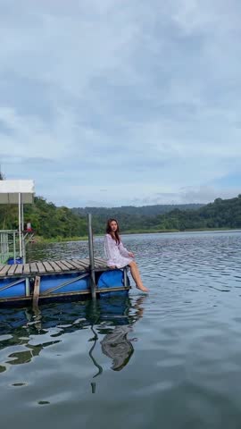 Beautiful woman wearing a dress is sitting relaxed on a wooden bridge while enjoying the natural beauty of the lake and hills. Woman vacationing in beautiful natural tourism.