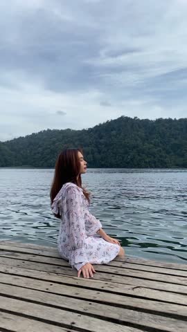Beautiful woman wearing a dress is sitting relaxed on a wooden bridge while enjoying the natural beauty of the lake and hills. Woman vacationing in beautiful natural tourism.