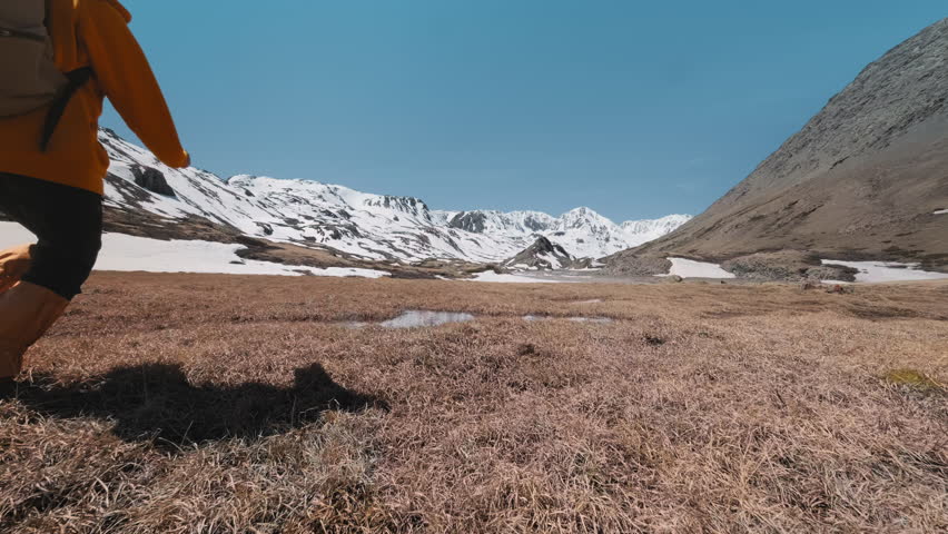 Couple or friends or tourists with backpacks or active hikers jumps over the water against the background of snowy mountains. People in colored clothes walks, runs and jumps in nature.