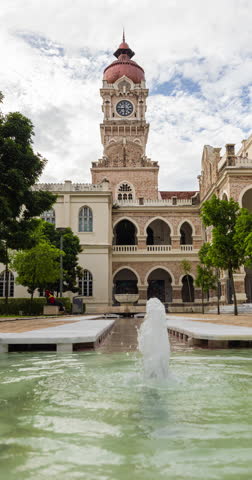 Vertical time lapse view of the clock tower from behind the Merdeka Square and near the river of life. Small water fountain behind the Sultan Abdul Samad Building in Kuala Lumpur, Malaysia, Timelapse