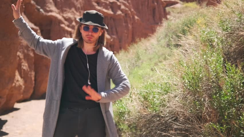 Long-haired guy in hat and casual clothes dancing and waves hands among the sandy desert mountains. Slow motion.