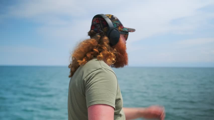 Bearded man in black glasses singing along to music on the phone dancing and having fun on a tropical beach.