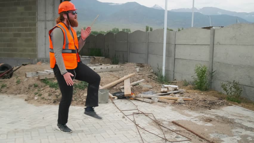 A charismatic builder dancing with robotic movements in slow motion, dressed in stylish orange work clothes and a hard hat, rests during a break against the backdrop of debris at a construction site.