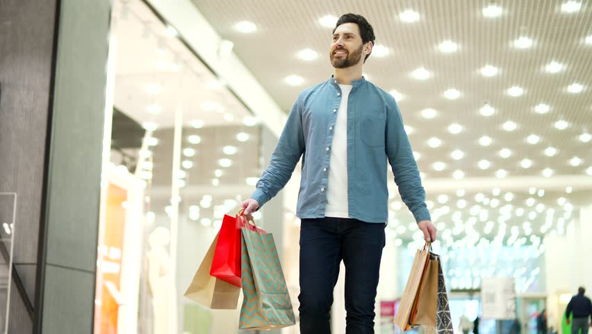 Happy satisfied handsome man walking in shopping center mall between shops store. Delighted male with shopping or gift packages bag on sale or discount day or black friday indoor plaza 