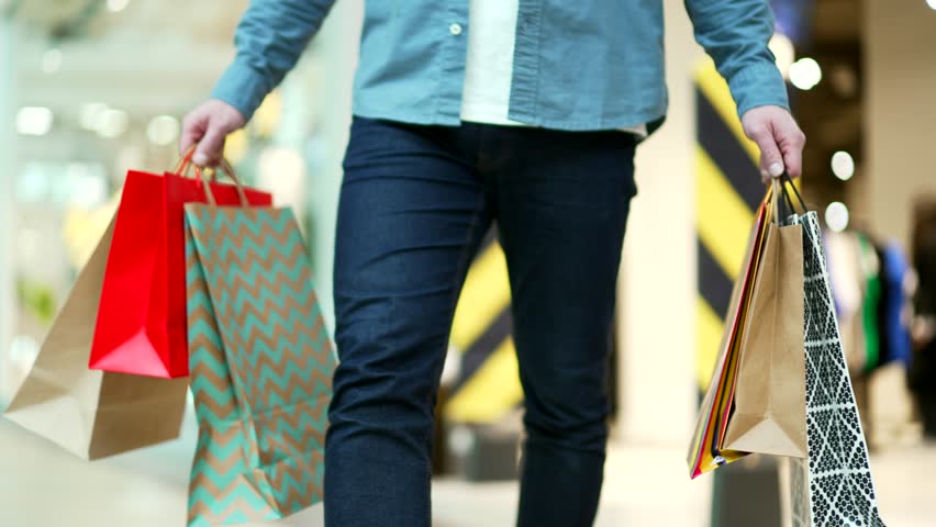 close up man walking in shopping center mall between shops. Unrecognizable shopaholic guy going at store Delighted male with gift packages bag on sale or discount day or black friday indoor plaza