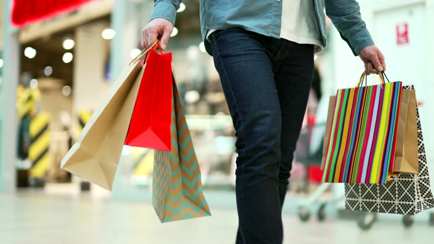 close up man walking in shopping center mall between shops. Unrecognizable shopaholic guy going at store Delighted male with gift packages bag on sale or discount day or black friday indoor plaza