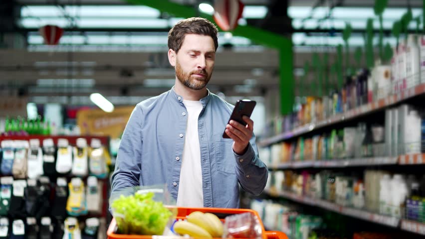 Handsome middle aged man buyer customer checking shopping list using smartphone, purchasing in grocery store in supermarket male shopper pick use mobile phone in hypermarket or food market deli