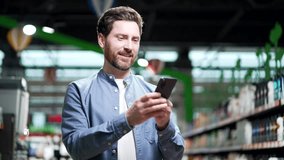 Happy adult man uses mobile phone in a supermarket while standing between the rows. Indoor Male typing text message, chatting browsing in smartphone in grocery store food market hypermarket or mall - Powered by Shutterstock - Get 15% off with code: PIKWIZARD15