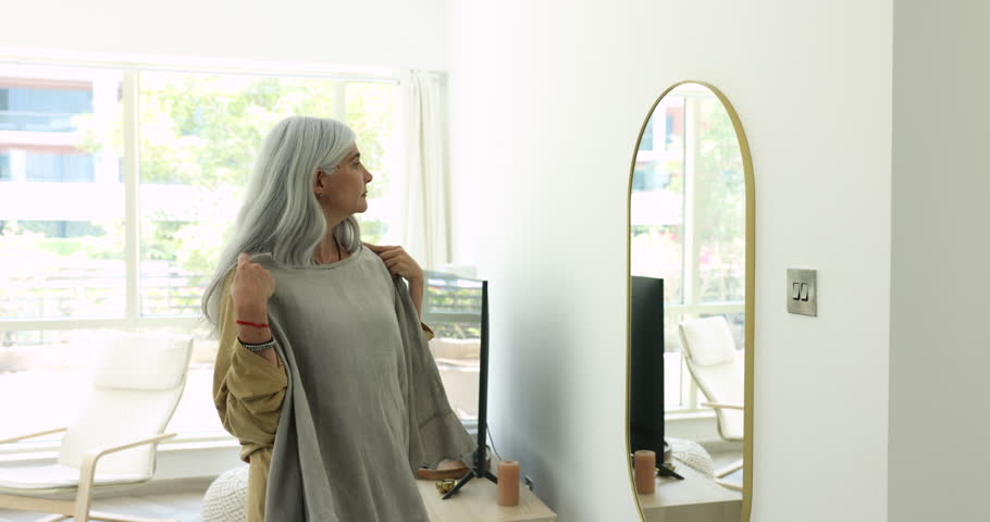 Stylish grey haired senior Latin woman trying on trendy oversized sweater, looking in home mirror, caring for style, fashion, outfit, choosing cloth for wearing, checking purchase from webstore