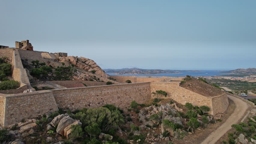 A drone view captures the ancient fortress walls and structures perched atop a rocky hillside in Sardinia, Italy. The blue waters of the Mediterranean Sea stretch out in the distance,