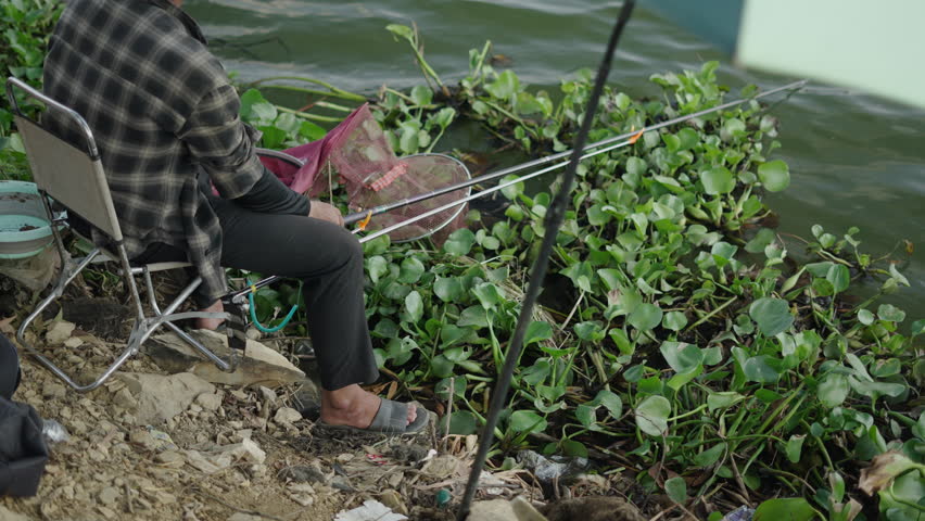 young fisherman holds the rod from him sitting on a chair on the rocks and vegetation in tri an lake vietnam