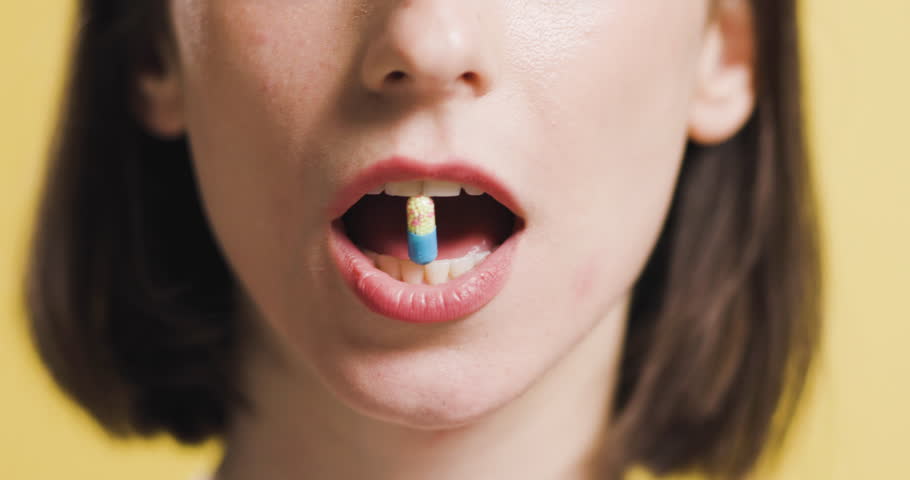 A close-up image of a Caucasian woman smiling as she holds a multicolored capsule between her teeth, swallowing, showcasing healthcare and medication.