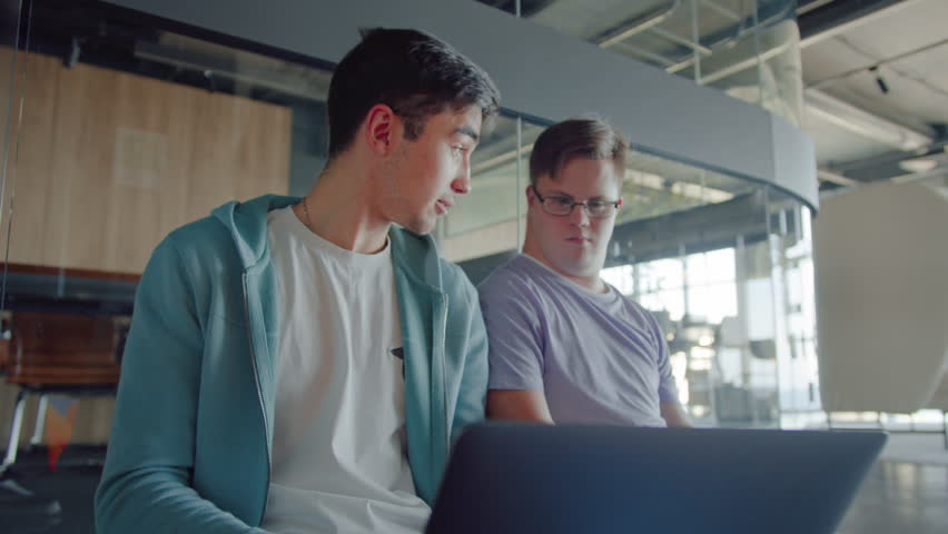Low angle view on caucasian person with down syndrome listening carefully to teammate with pc on laps. Handsome guy giving instructions about project to colleague in sophisticated office.
