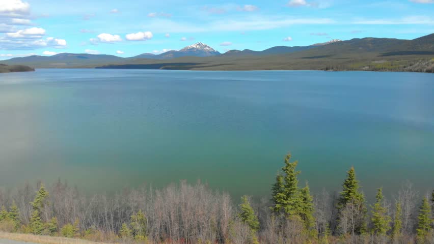 Stunning aerial view of a serene lake in Yellowstone National Park, surrounded by lush forests and snow-capped mountains under a clear blue sky, capturing nature’s tranquility.
