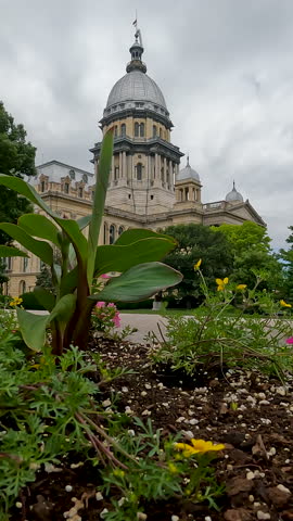 Views of the Illinois State Capitol Building in Springfield, IL, USA as viewed through a blooming floral garden. Dramatic cloudscape and green leafy trees surround the scene.