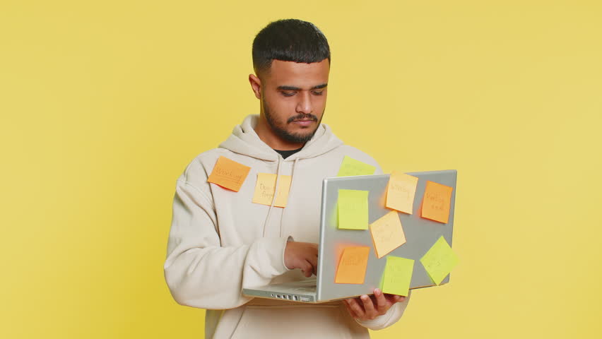 Exhausted freelancer Indian man with pasted sticker notes use laptop computer having concentration problem creative crisis. Nervous Arabian guy confused by big routine work loading yellow background