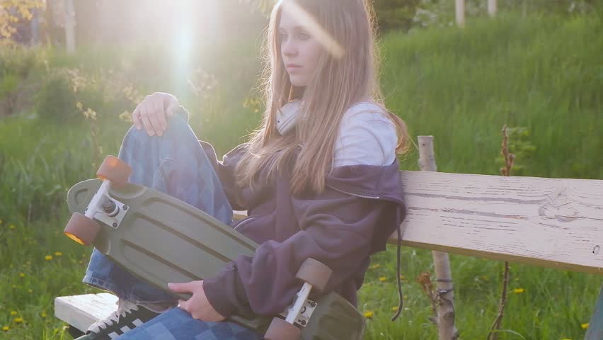 A beautiful cool teenage girl with Wearing headphones holds a skateboard and sits on an outdoor bench.