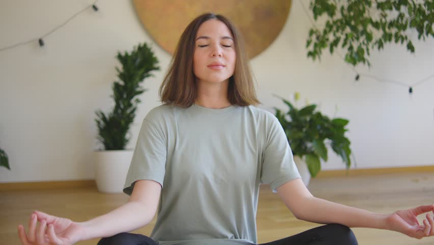 A young woman practices yoga indoors, focusing on wellness, relaxation, and spirituality. Her serene environment with plants and natural light promotes a healthy lifestyle and stress relief